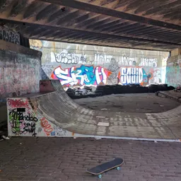 A skateboard rests in front of a worn ramp under a bridge covered in graffiti.