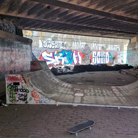 A skateboard rests in front of a worn ramp under a bridge covered in graffiti.